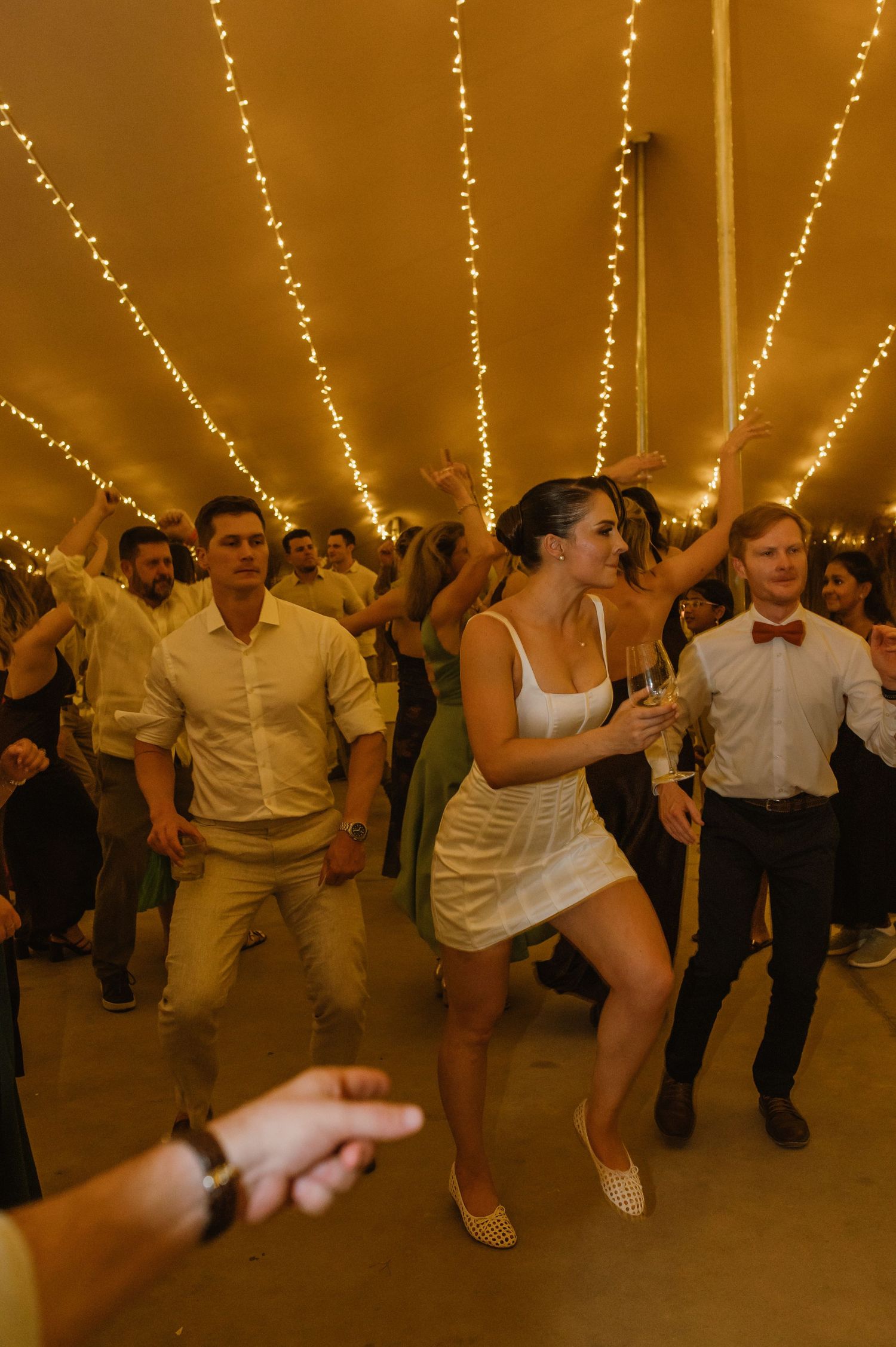 People dancing and celebrating at a wedding reception under warm string lights in a tent.