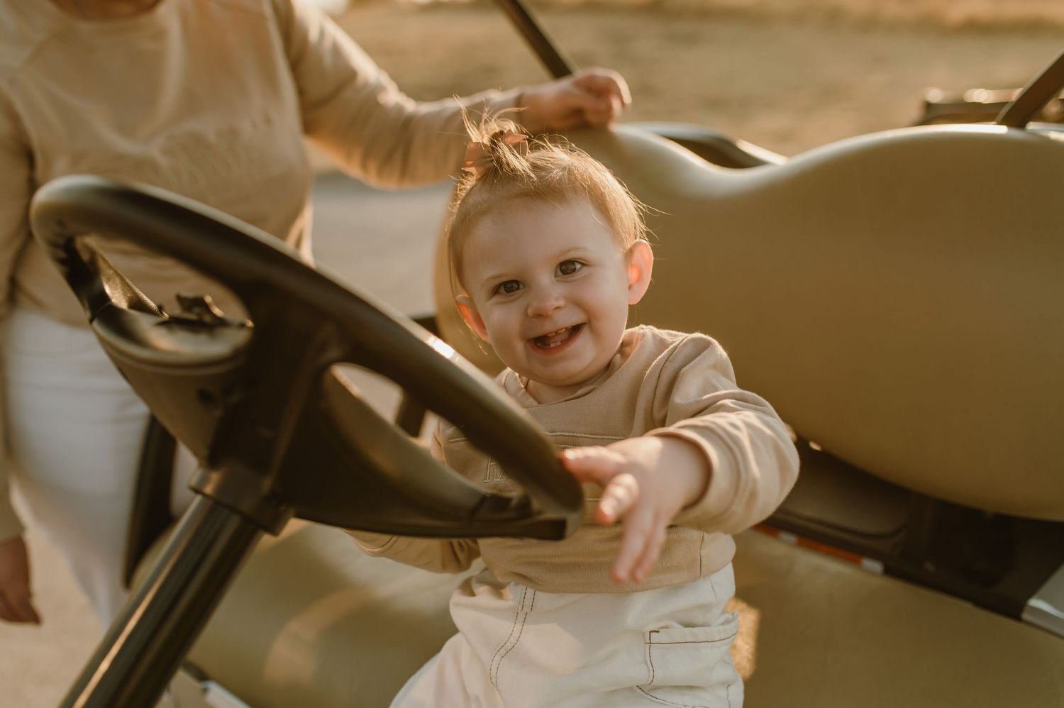 A toddler in a beige sweater smiles while playing at the steering wheel of a large vehicle.