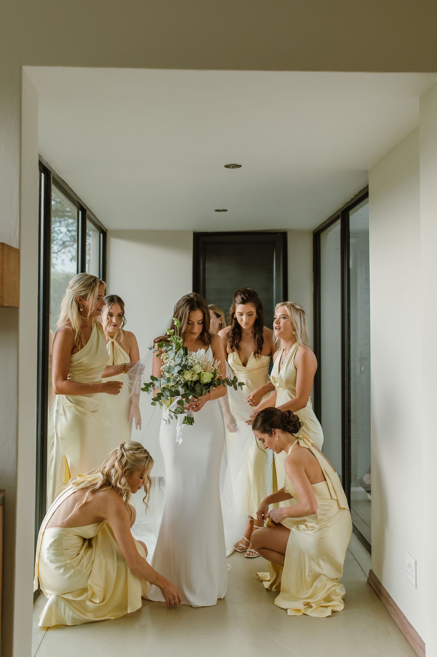 Bridesmaids in pale yellow dresses gather in a modern hallway with floor-to-ceiling windows.