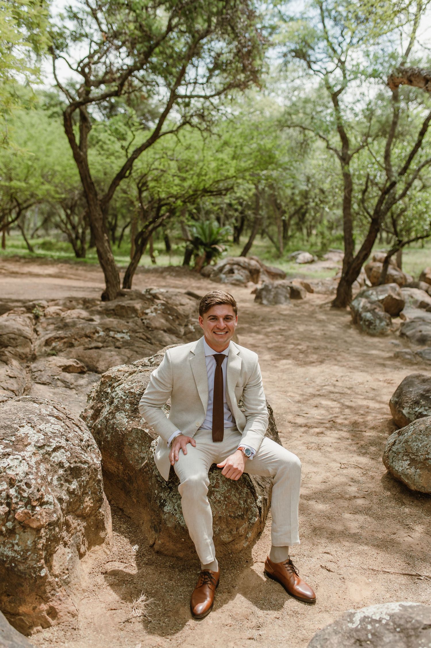 A groomsman in a light suit and tie sits casually on a large rock in a wooded outdoor setting.