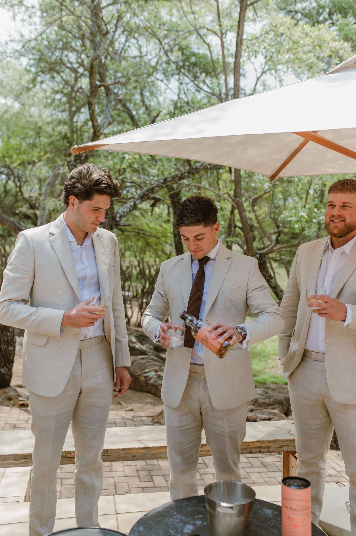 Groomsmen in beige suits share drinks together at an outdoor wedding venue.