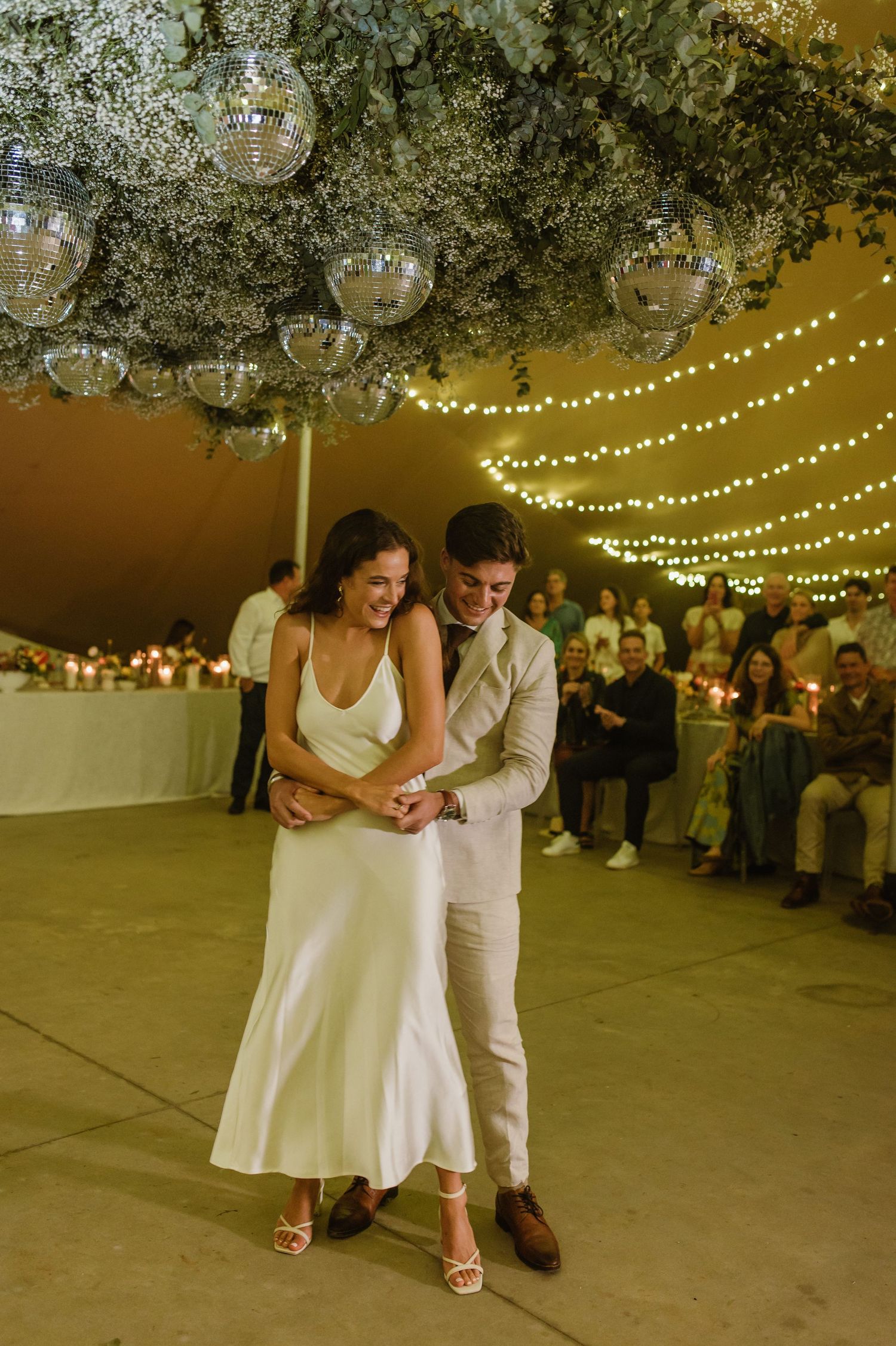 Couple shares their first dance under disco balls and string lights at an indoor wedding reception.