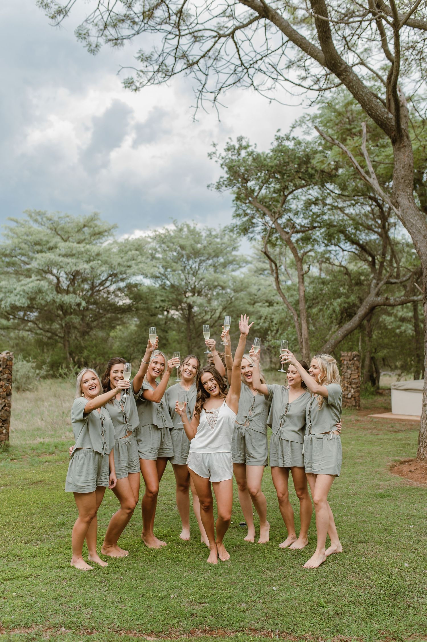 Group in coordinated sage outfits pose together with raised arms on a grassy lawn.