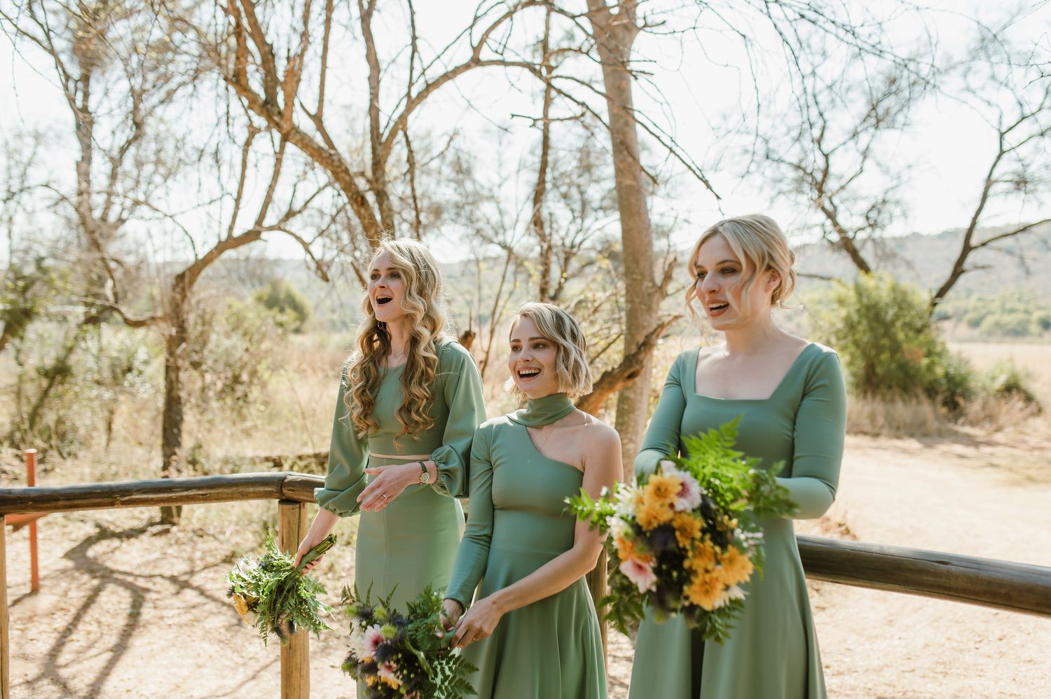 Wedding party in sage green dresses posing on wooden deck with floral bouquets.