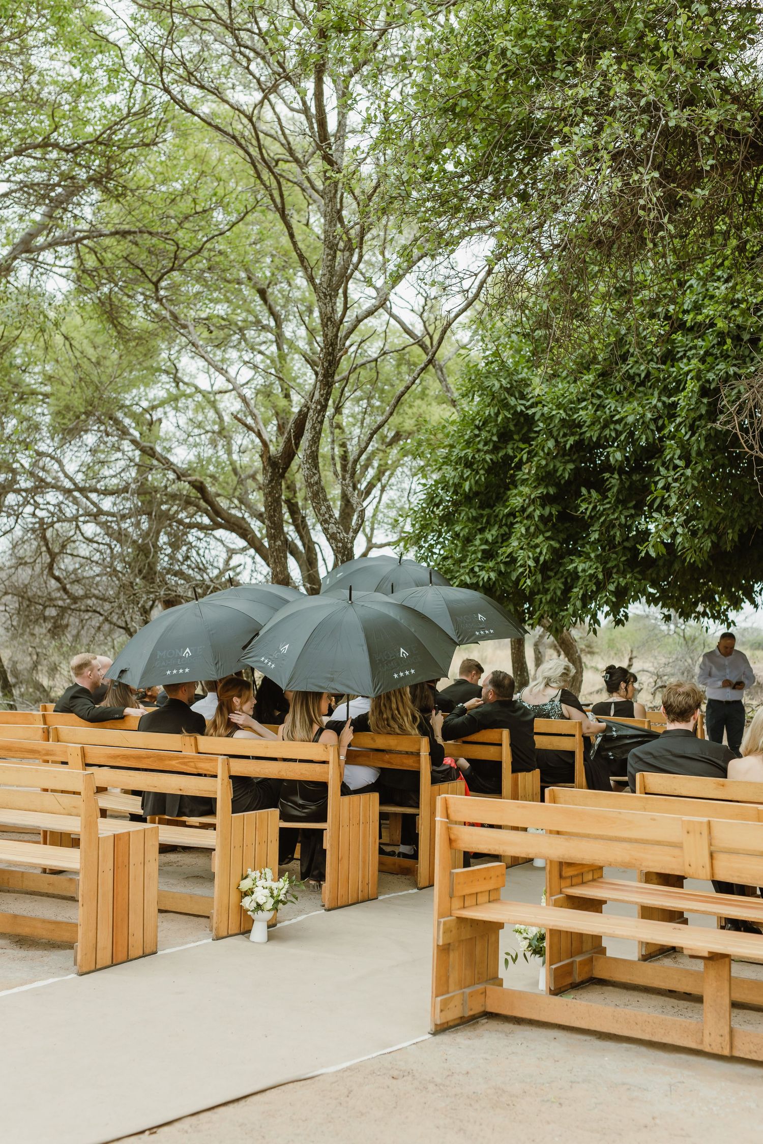 Wedding guests with black umbrellas seated on wooden benches under green trees.