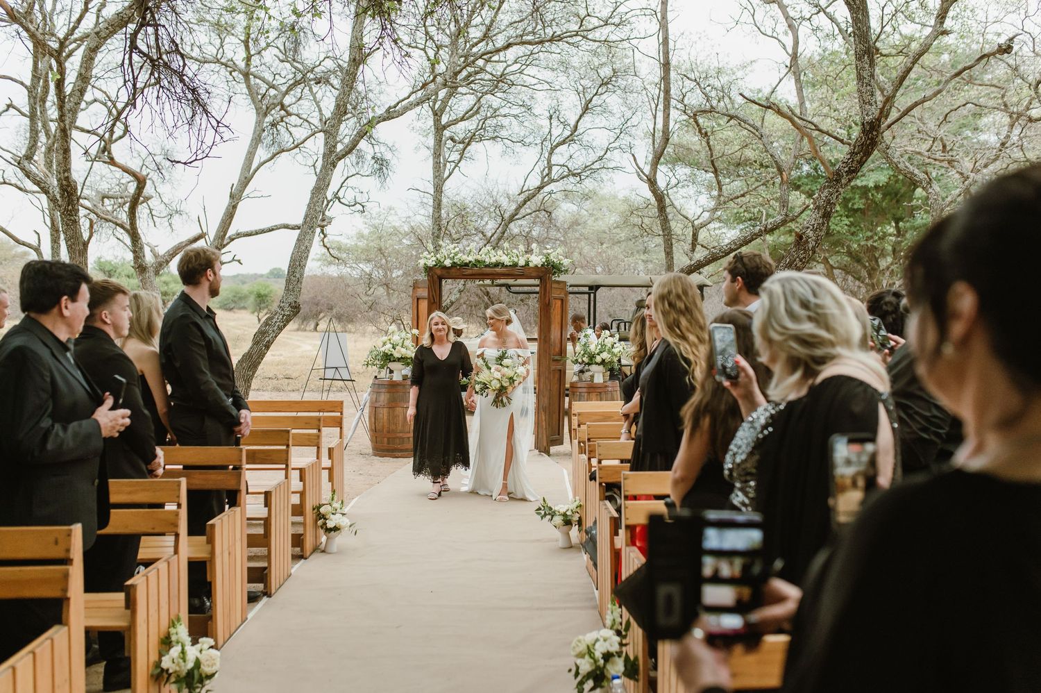 Wooden benches line a natural outdoor wedding venue under tree canopy.