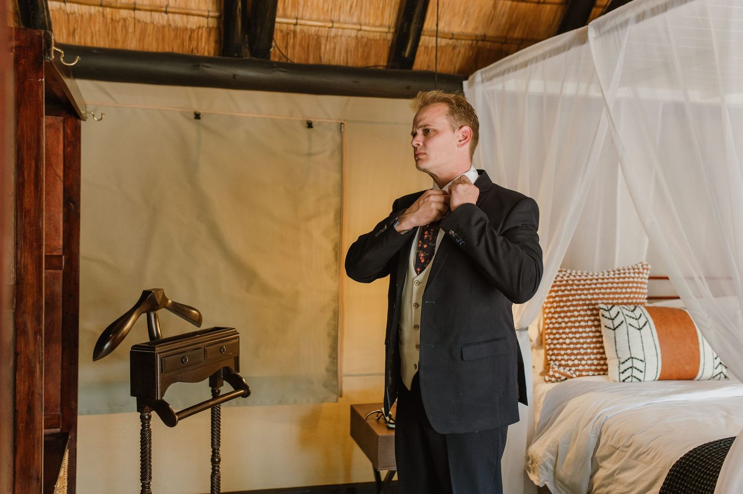 Person in formal attire adjusting their tie in a bedroom with thatched roof.