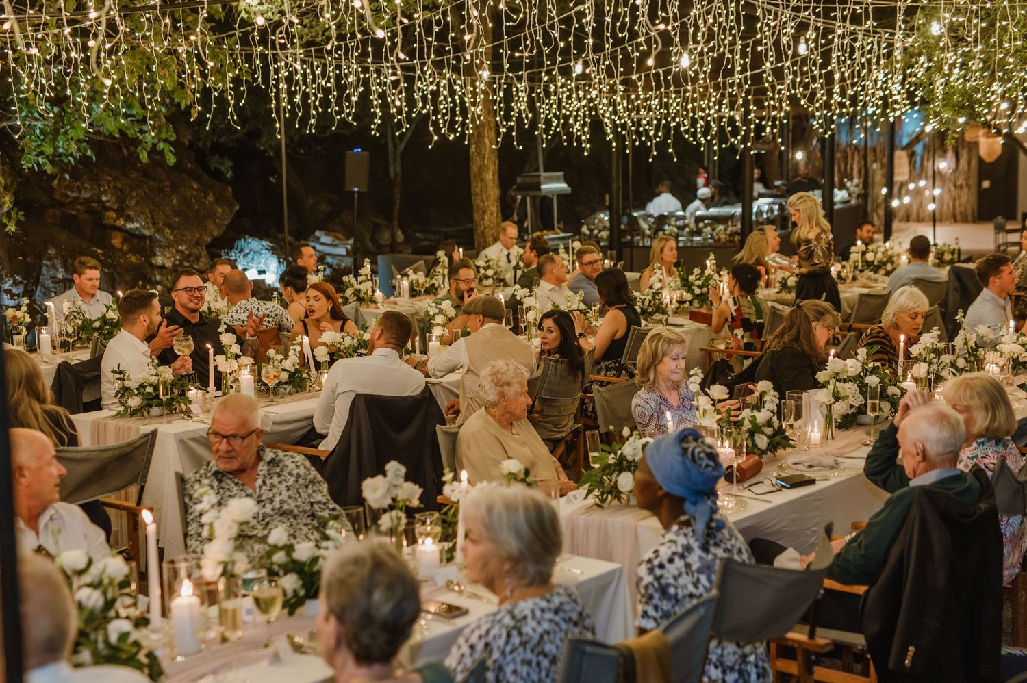 Panoramic view of wedding reception with guests dining under glowing string lights and natural canopy.