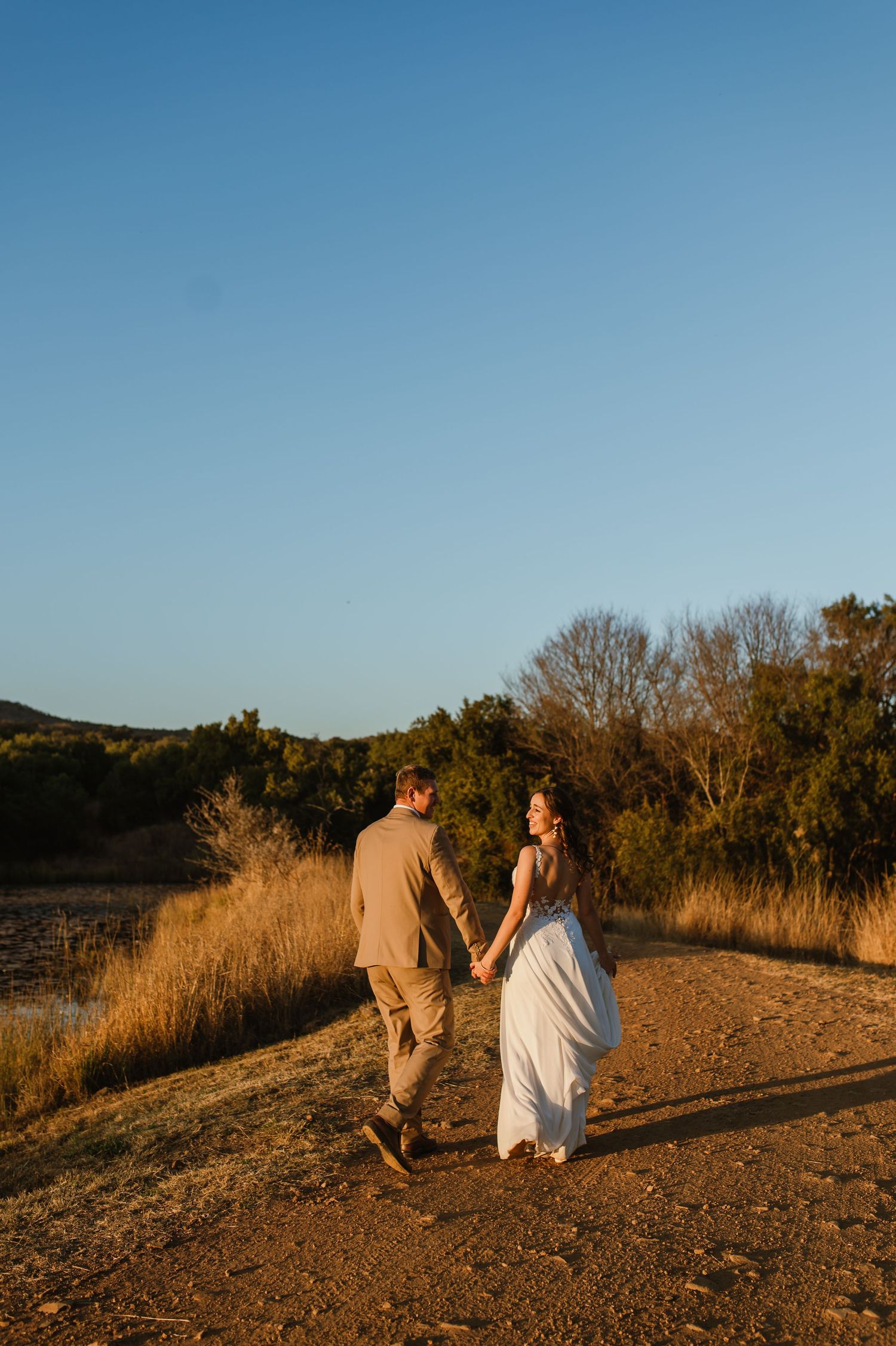 A couple walks together on a dirt path during golden hour with trees and brush in the background.