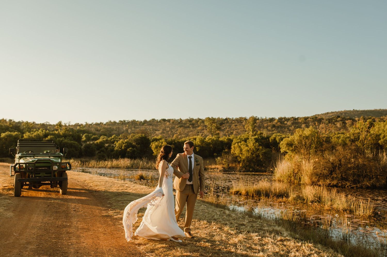 Wide landscape shot of a wedding party and vehicles on a dirt road at sunset.