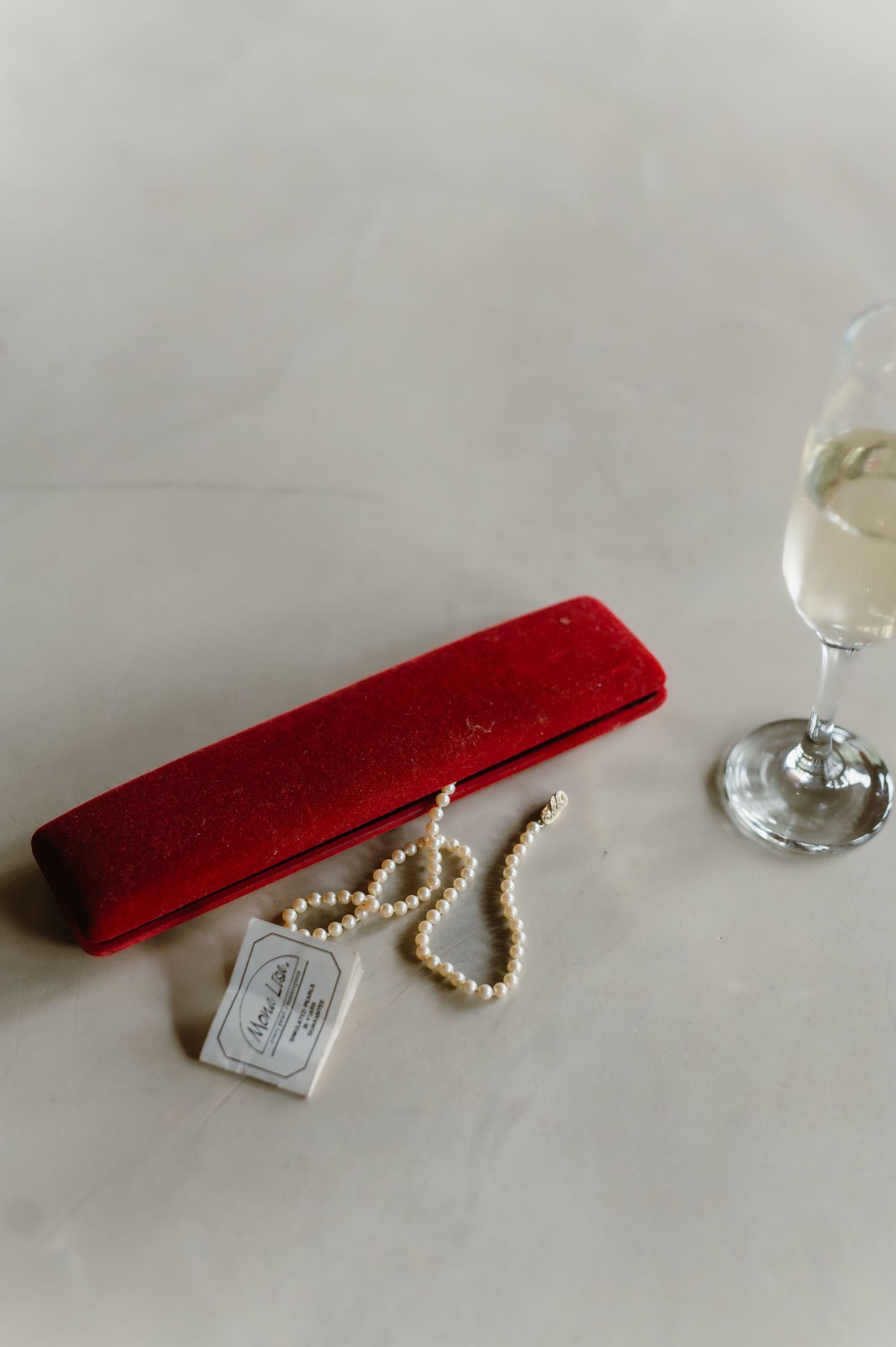 Pearl necklace with red velvet box and wine glass on white surface.