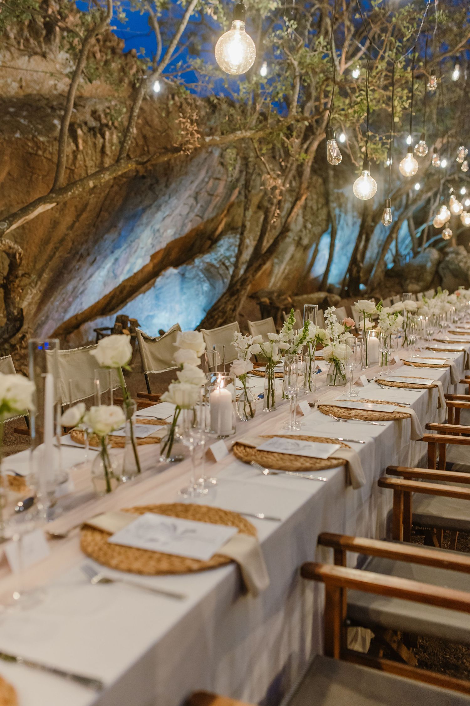 Rustic wedding reception tables set with woven placemats and white linens under natural tree canopy.