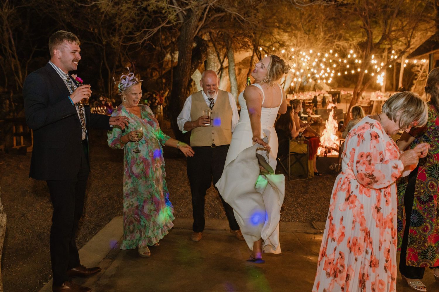 Wedding guests dance under twinkling lights at outdoor evening reception.