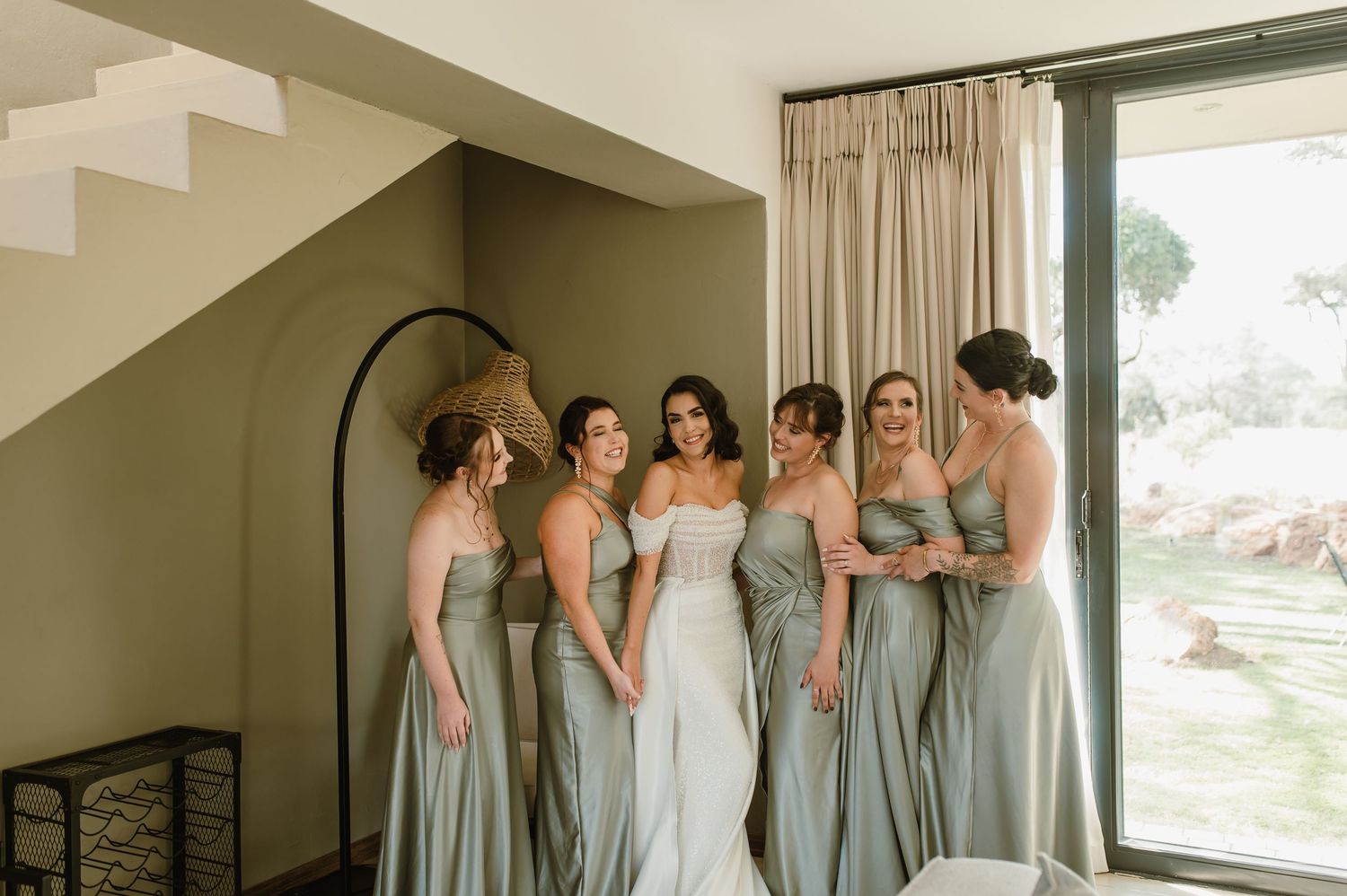 Bridal party in sage green dresses arranged in front of large windows and modern architecture.