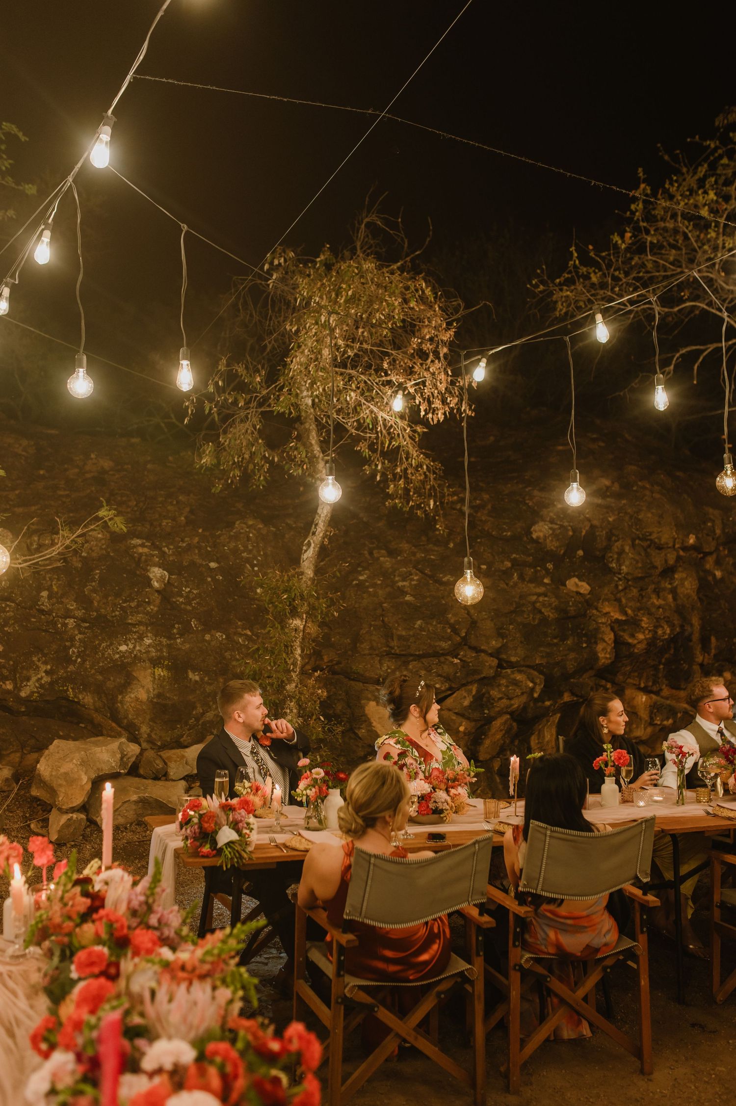 Romantic outdoor dinner setting with string lights and wooden tables arranged under trees at night.