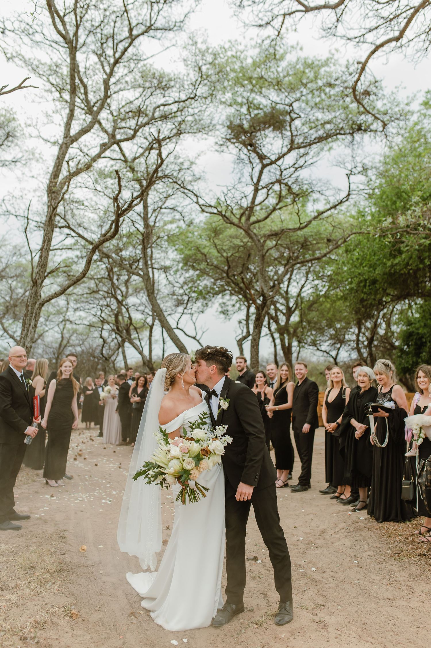 Romantic wedding moment captured on tree-lined path with guests in formal attire.