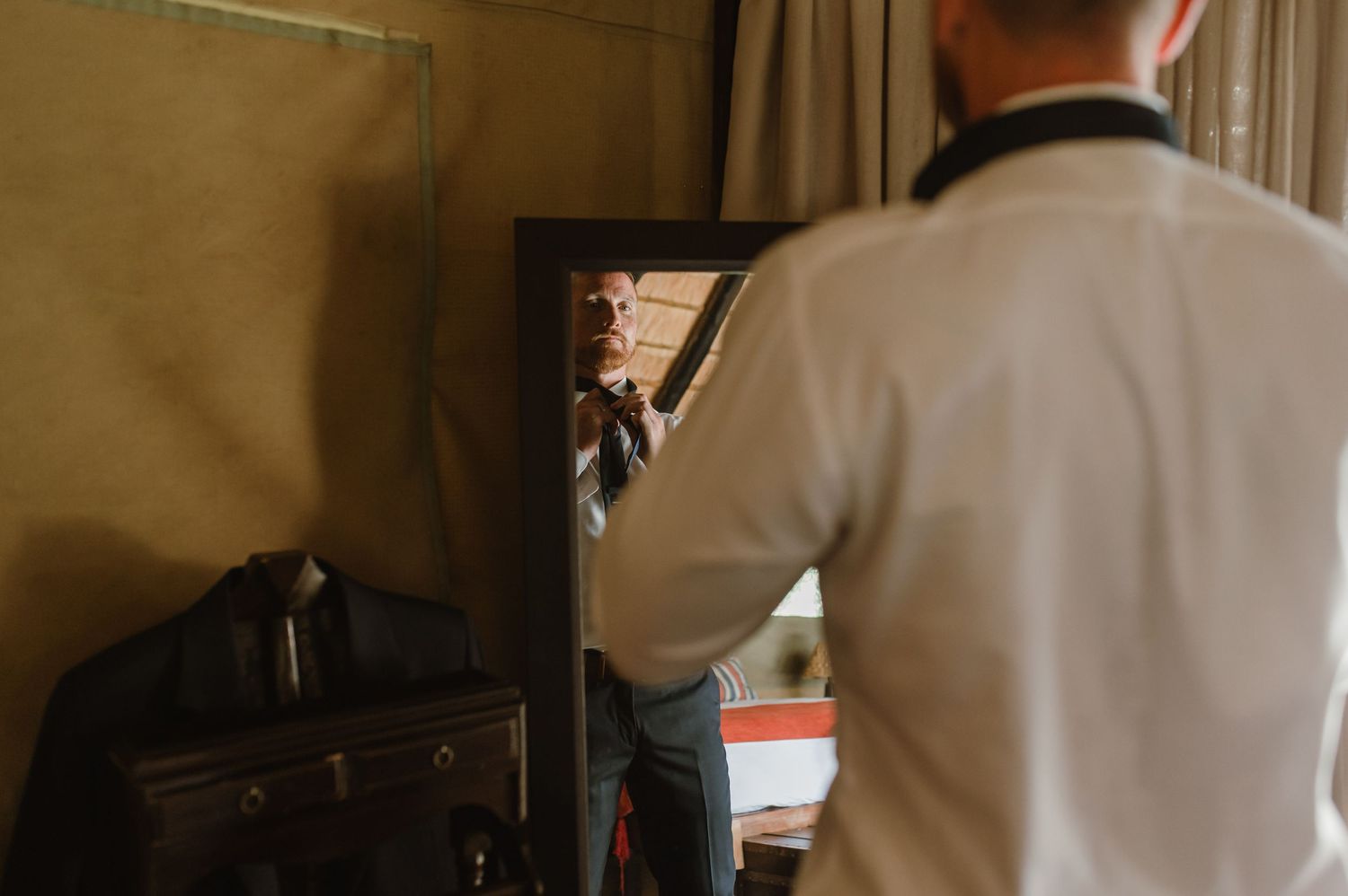 Men in formal black suits getting ready in a rustic room with wooden beams and thatched roof.