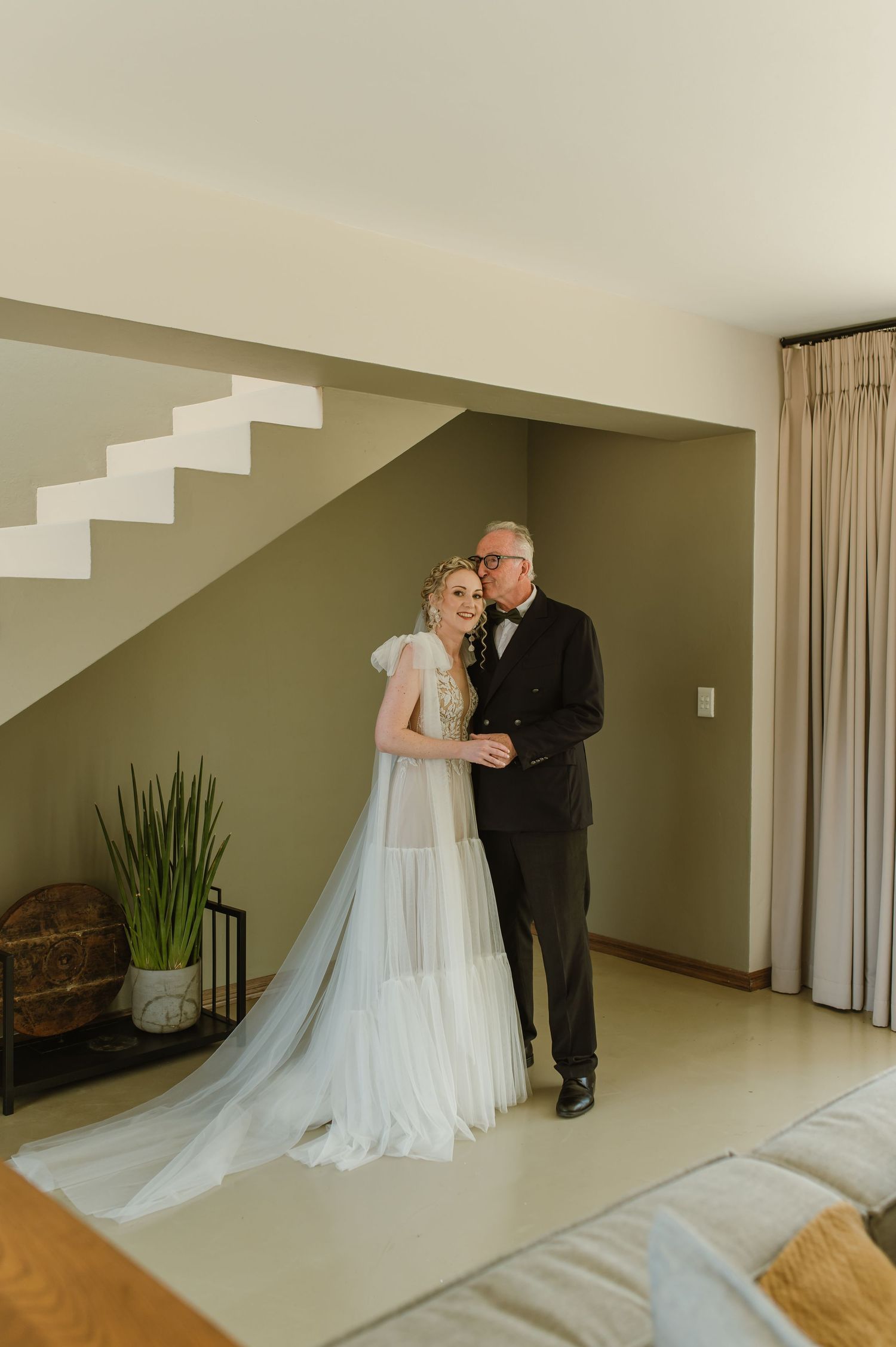 A wedding couple shares an intimate moment in a minimalist indoor setting with white walls and stairs.