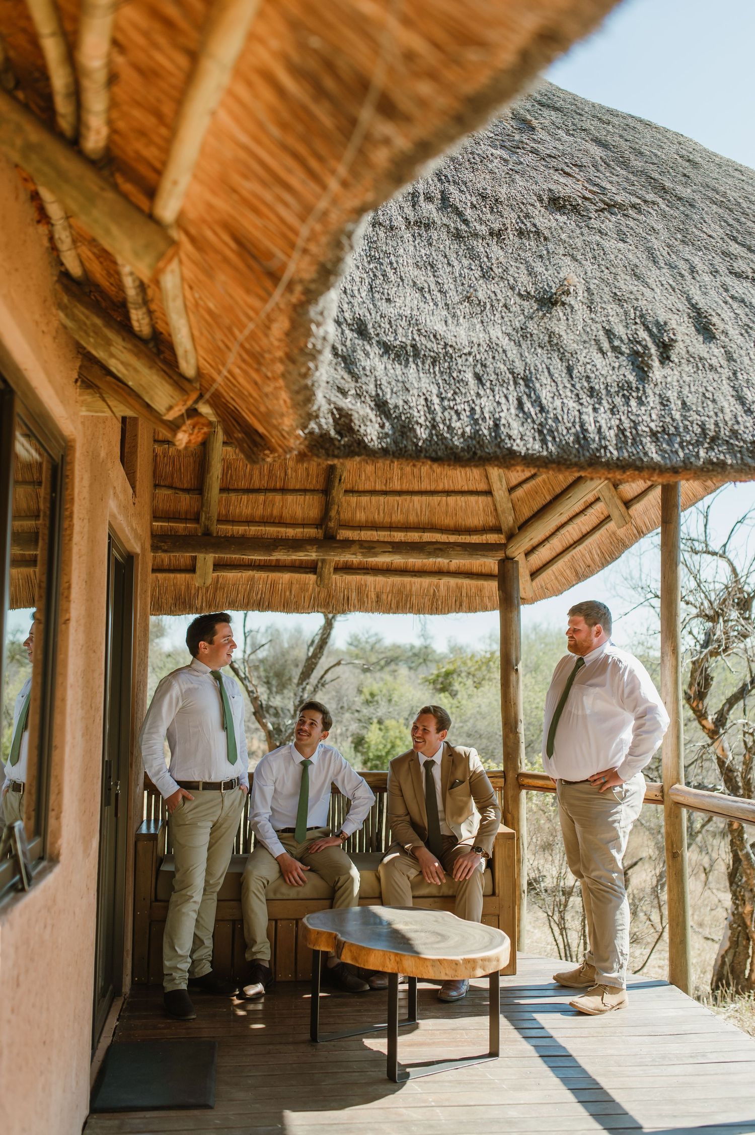 Wedding party members gathered under a thatched roof pavilion in safari setting.