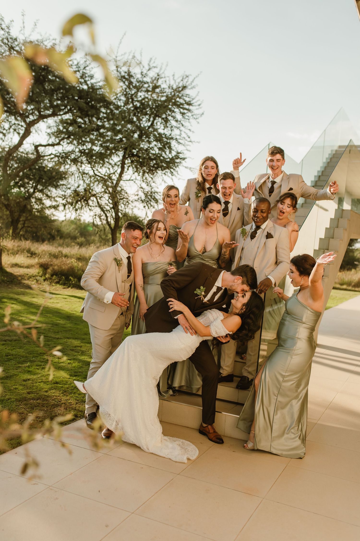 Wedding party poses playfully around white vehicle during outdoor celebration.