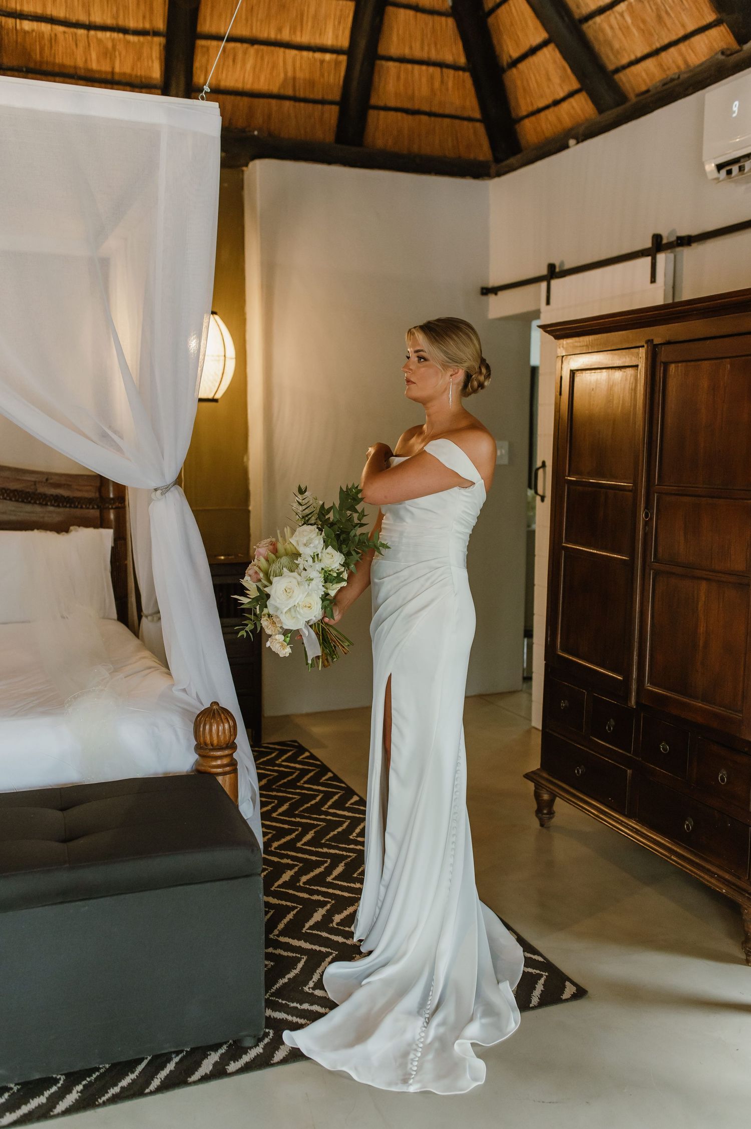 Bride in form-fitting white dress poses beside canopy bed in rustic room.