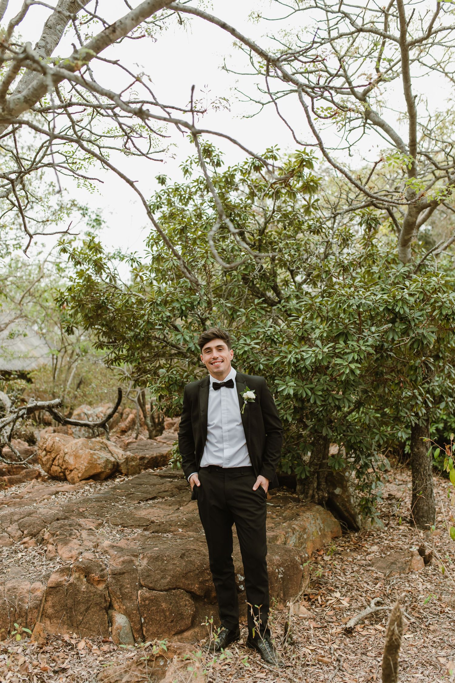 Groomsman in classic tuxedo poses among trees in natural outdoor setting.
