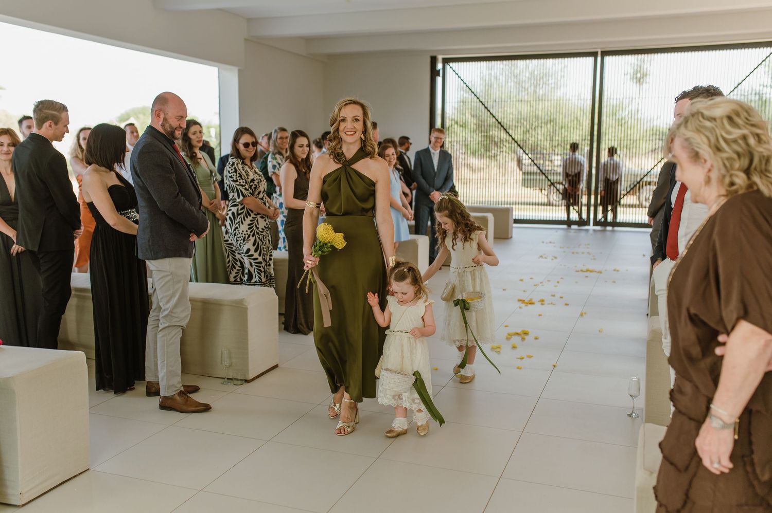 Wedding attendees line up along a bright minimalist hallway with floor-to-ceiling windows.