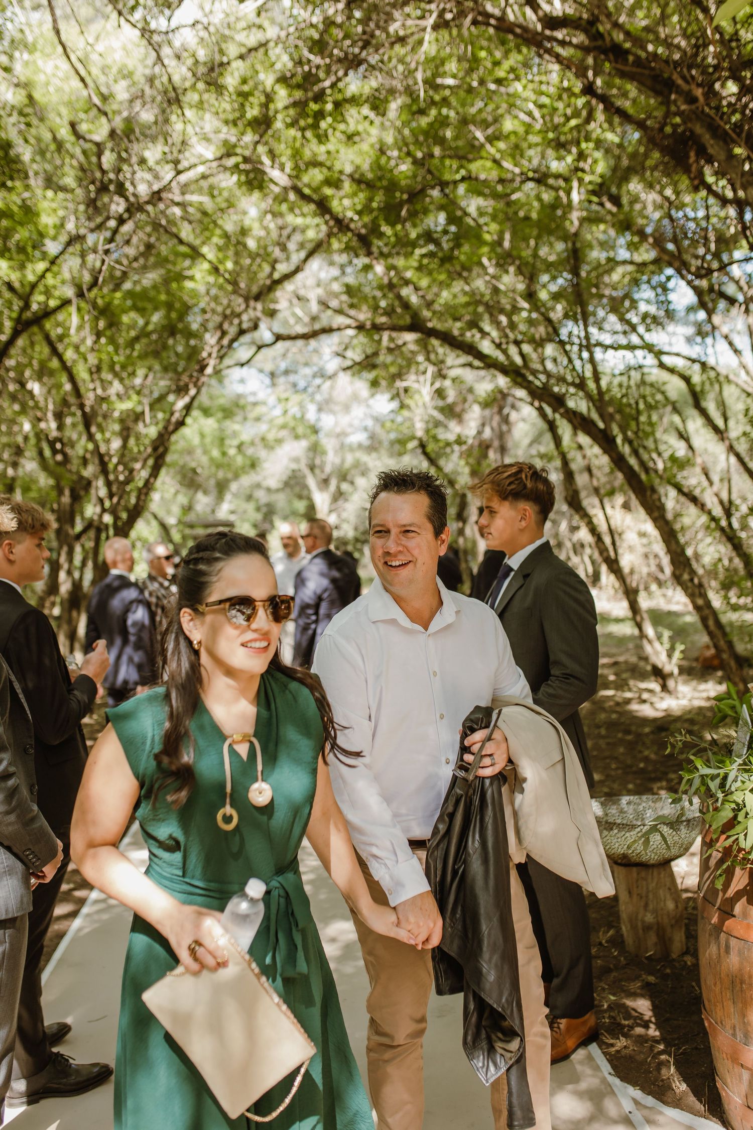 Wedding guests in casual attire socialize under a shaded tree canopy.