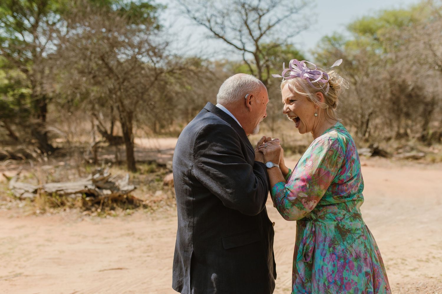 Couple in formal attire embracing on a dirt path surrounded by desert vegetation.
