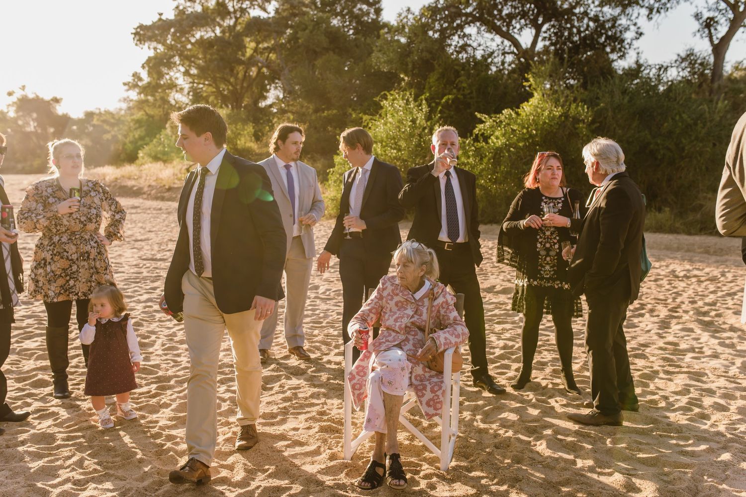 Large group of wedding guests mingling on sandy beach during golden hour.