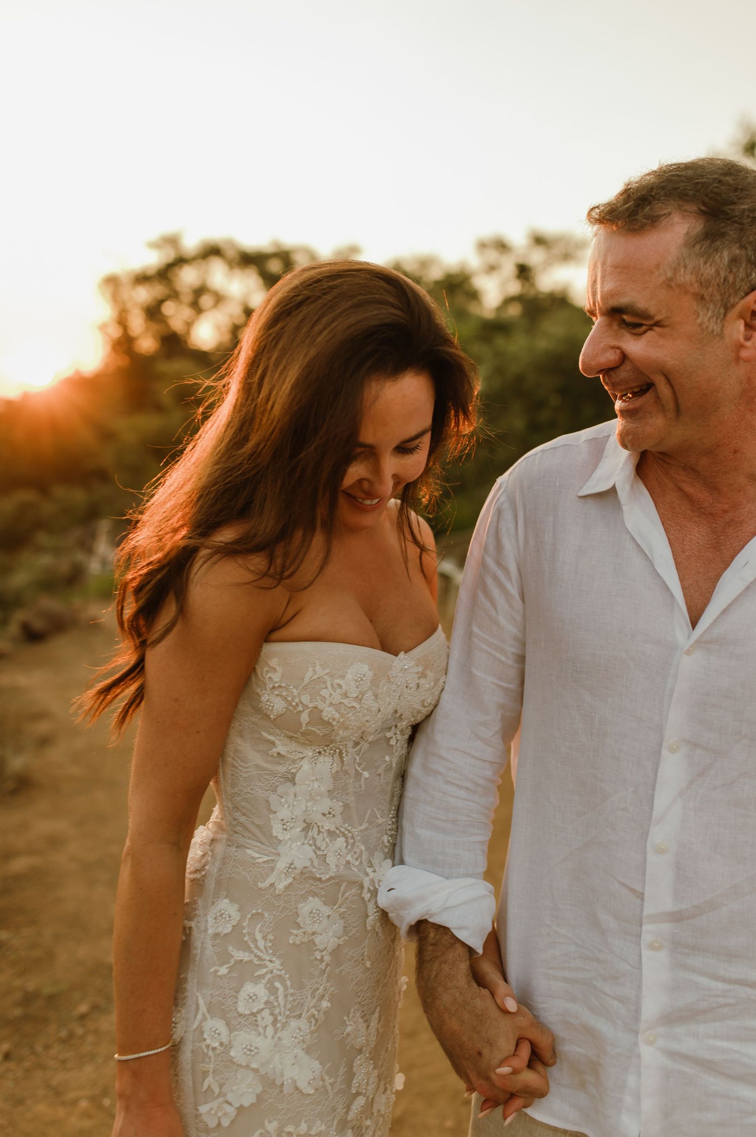Bride and groom laughing and holding hands while walking together in warm golden sunset light.