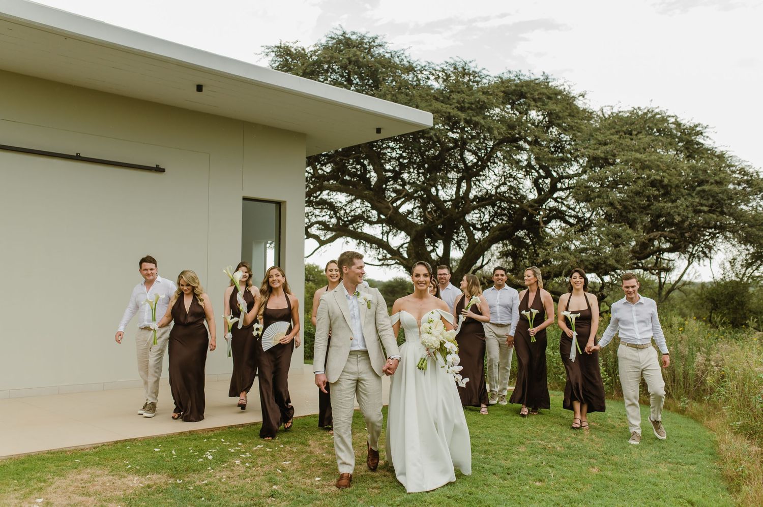 Wedding party stands in line against lush green field and modern white building backdrop at outdoor ceremony.