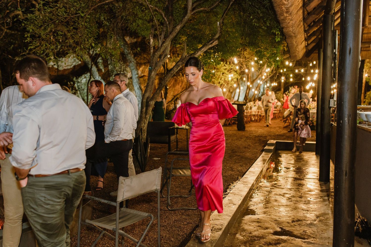 Woman in elegant red off-shoulder midi dress walks through outdoor wedding reception at night.