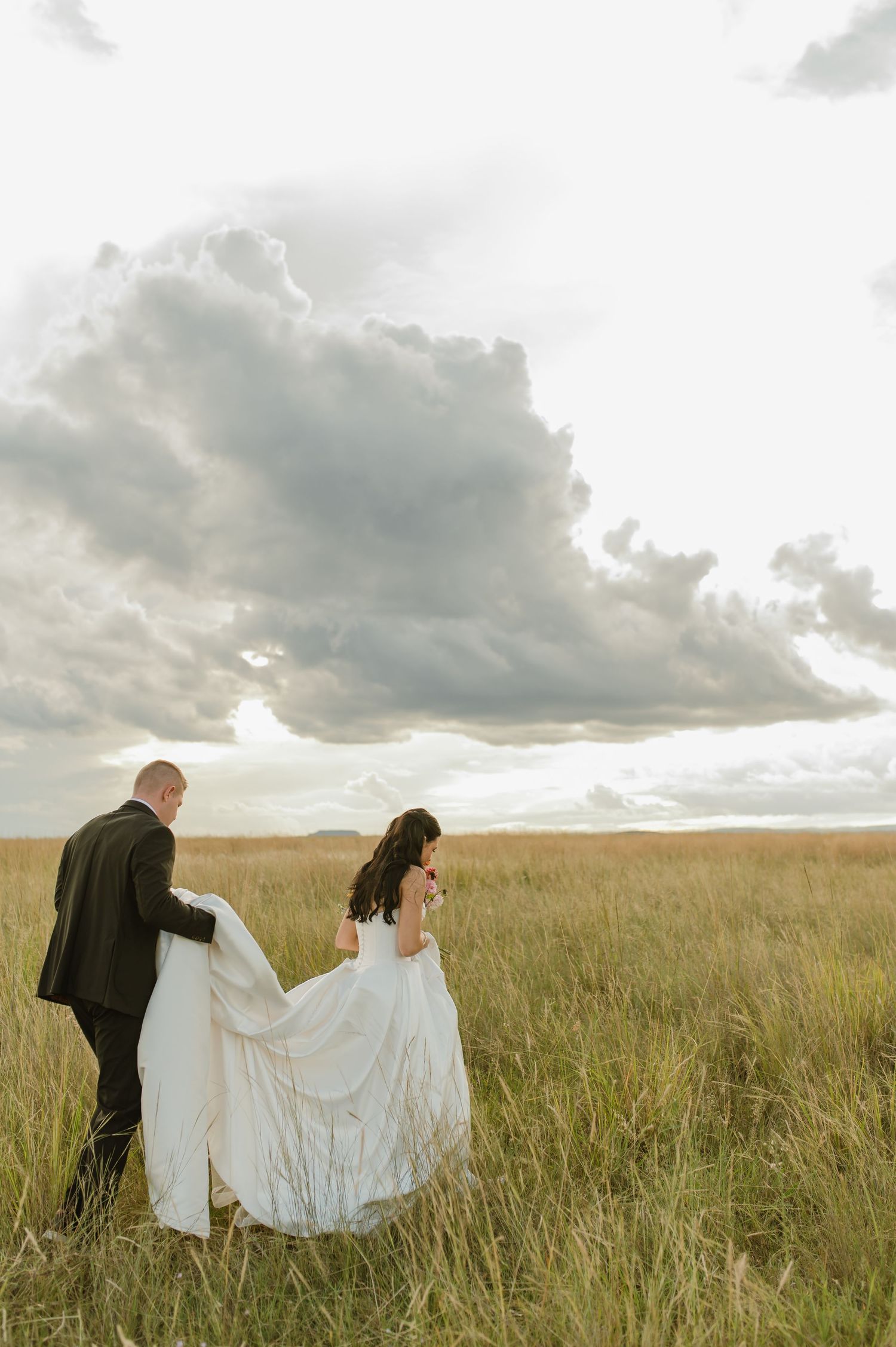 Bride and groom walk through a golden grass field under dramatic cloudy skies during their wedding portraits.