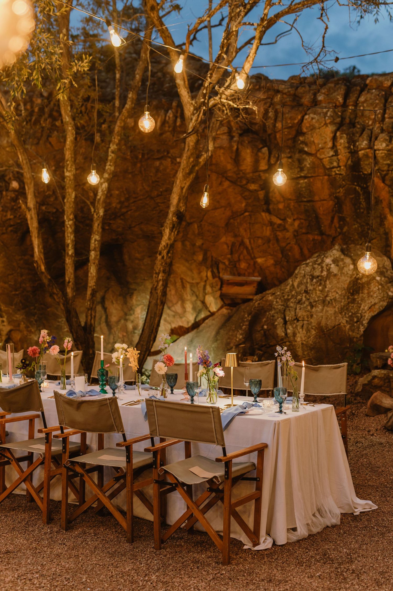 Elegant outdoor wedding reception table with string lights, candles, and colorful flowers at dusk.