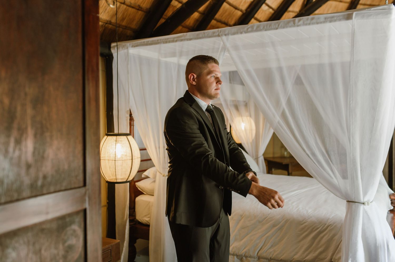 A groom in a dark suit stands beside a canopy bed with white drapes in a rustic tropical villa bedroom.