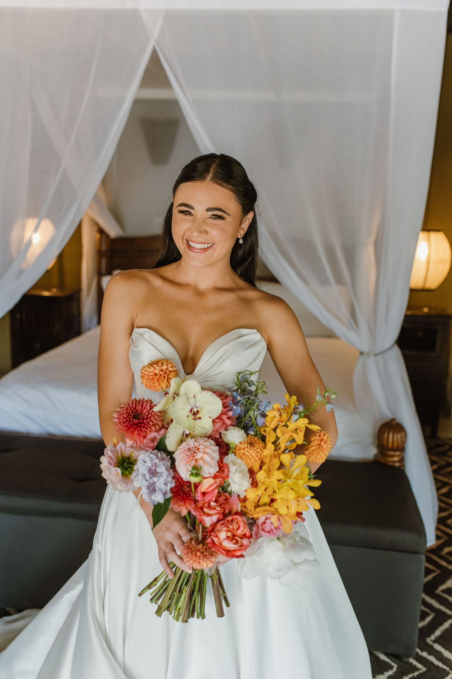 Smiling bride in a strapless white gown holding a colorful bridal bouquet with dahlias and lilies indoors.