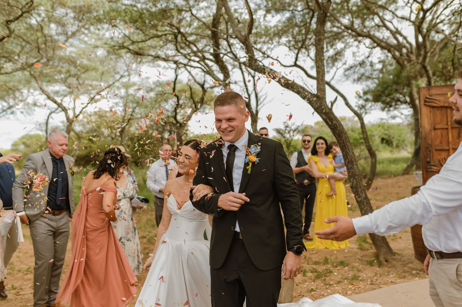 Newlyweds walk through confetti shower surrounded by cheering guests after outdoor ceremony.