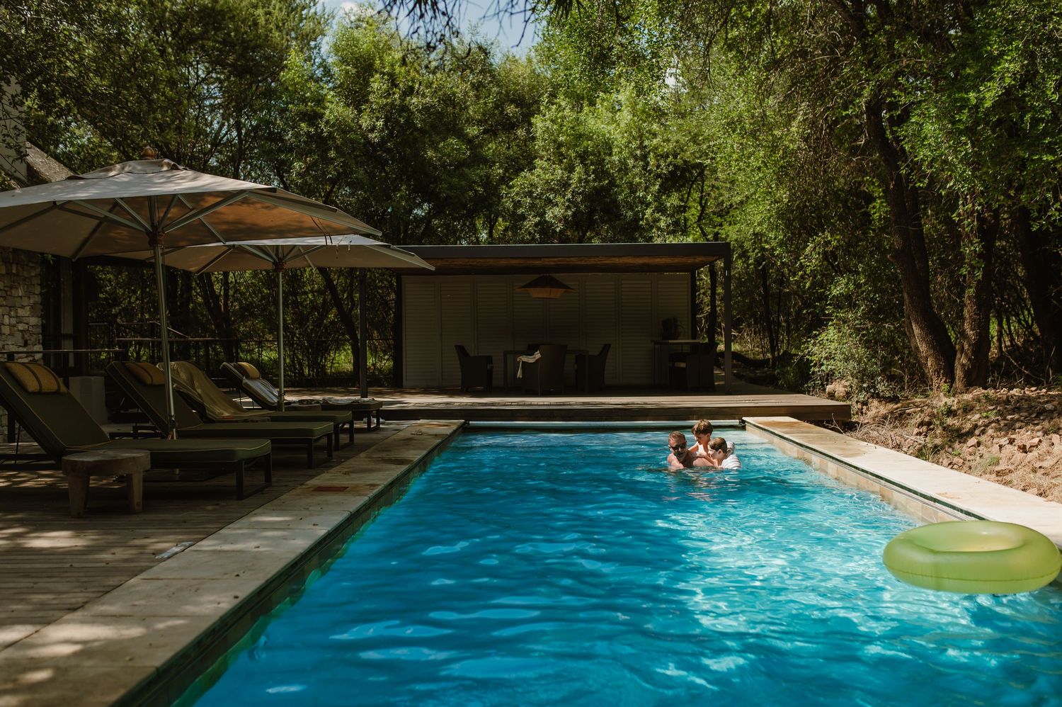 Guests swimming in a luxury safari lodge pool surrounded by lush trees and lounge chairs.