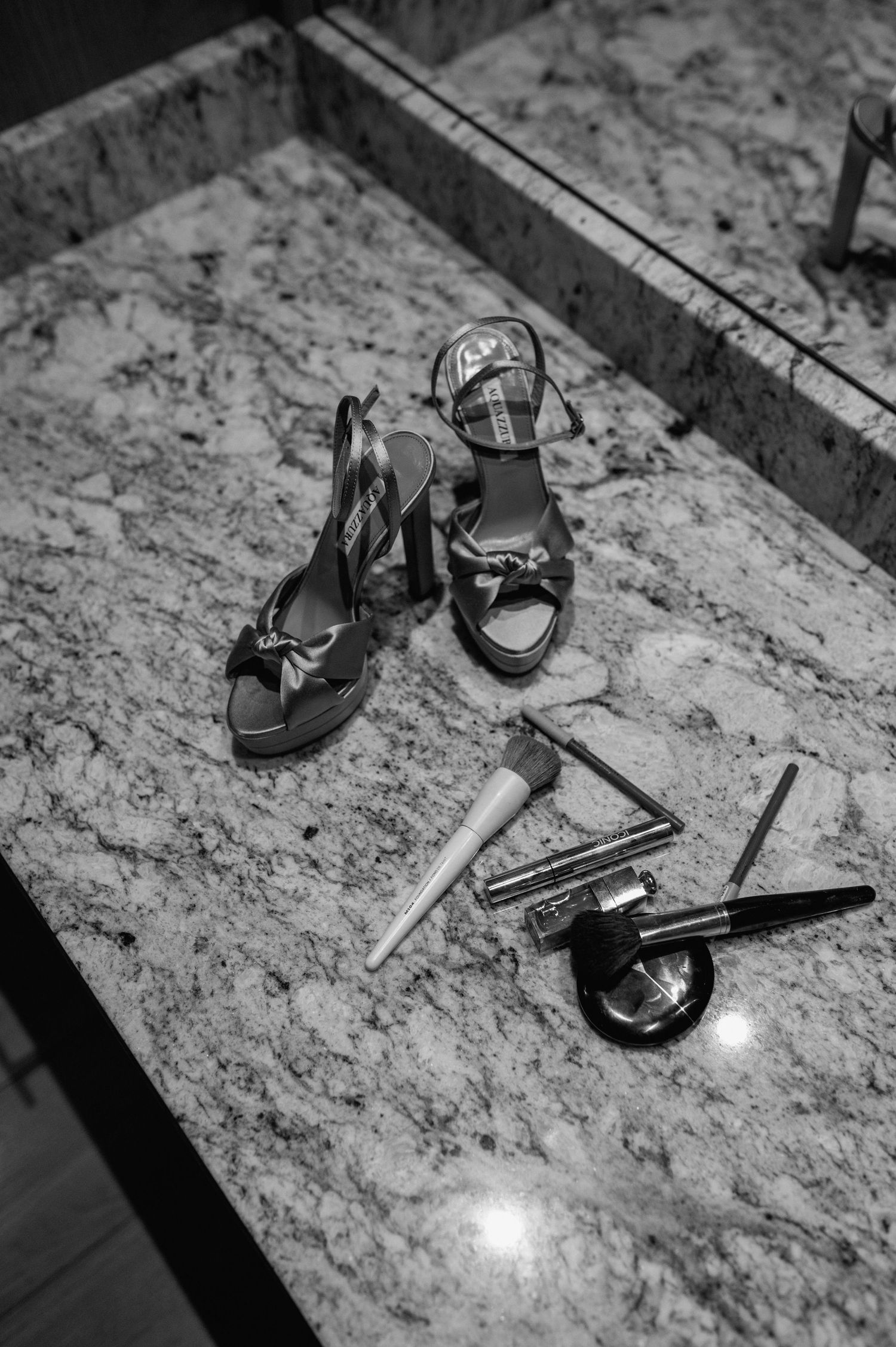 Black and white flat lay of bridal heels and makeup accessories on marble surface.