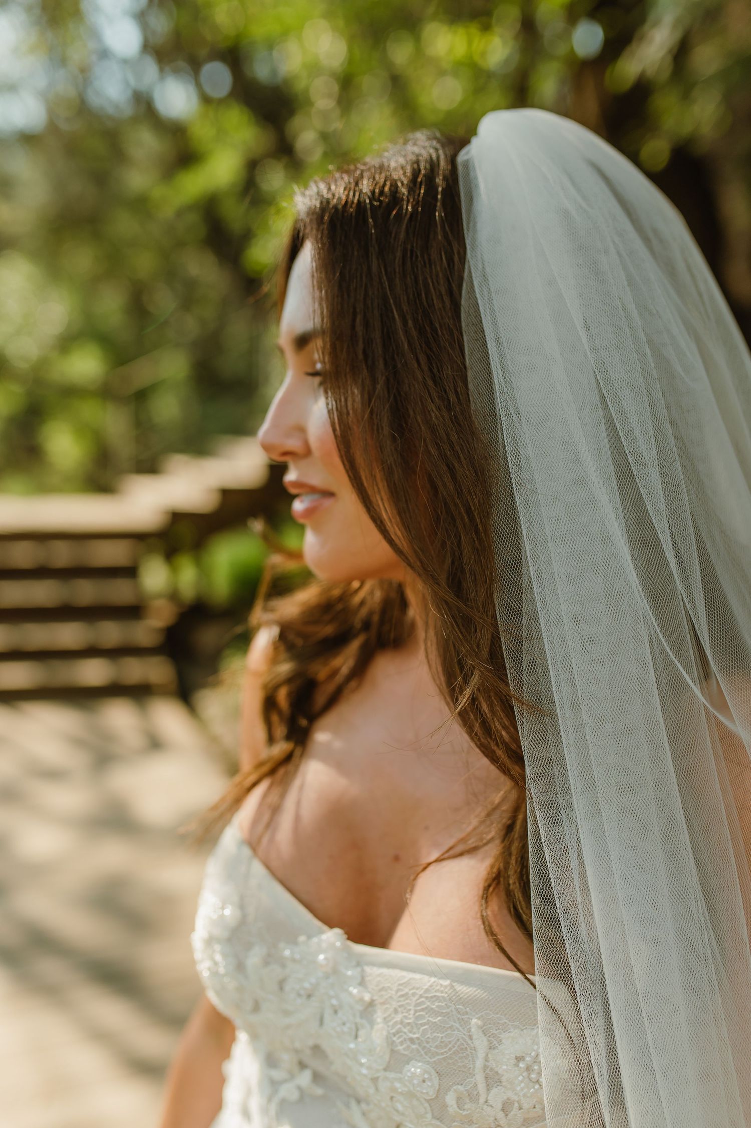 Beautiful bride with long brown hair wearing white veil in dappled sunlight outdoors.