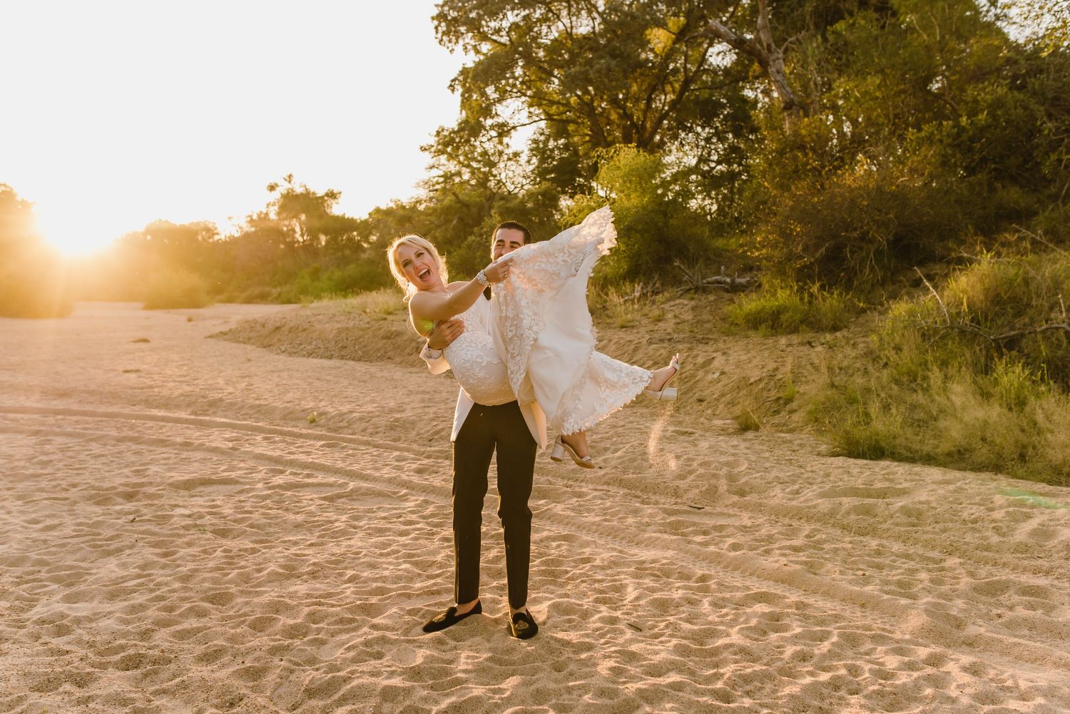 Playful wedding moment captured on beach as groom lifts bride during sunset.