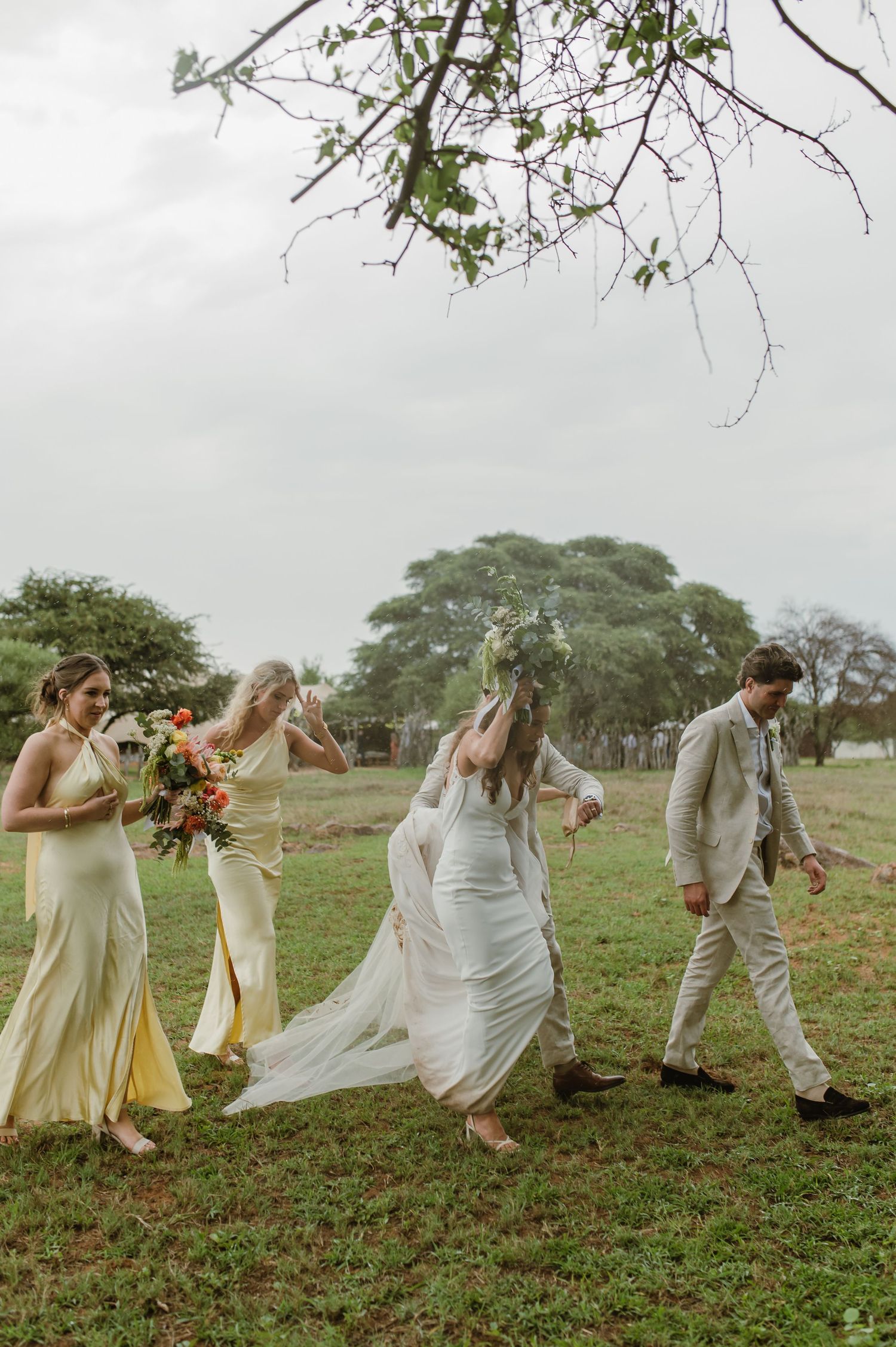 Wedding party members run and dance across a grassy field in celebratory poses.
