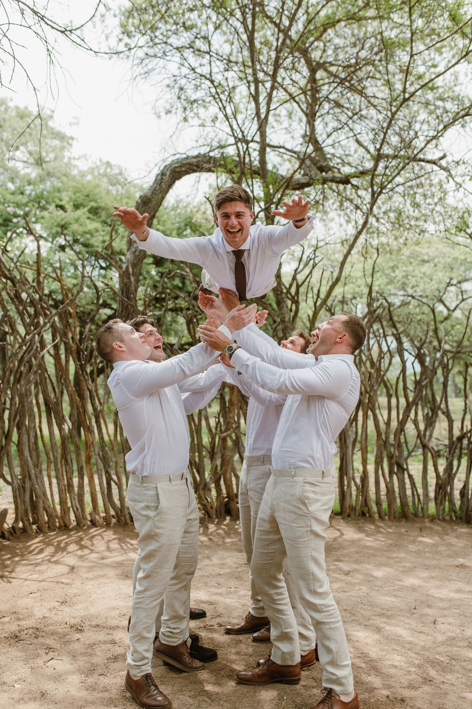 Wedding party members in light suits celebrate with playful poses in a natural setting.