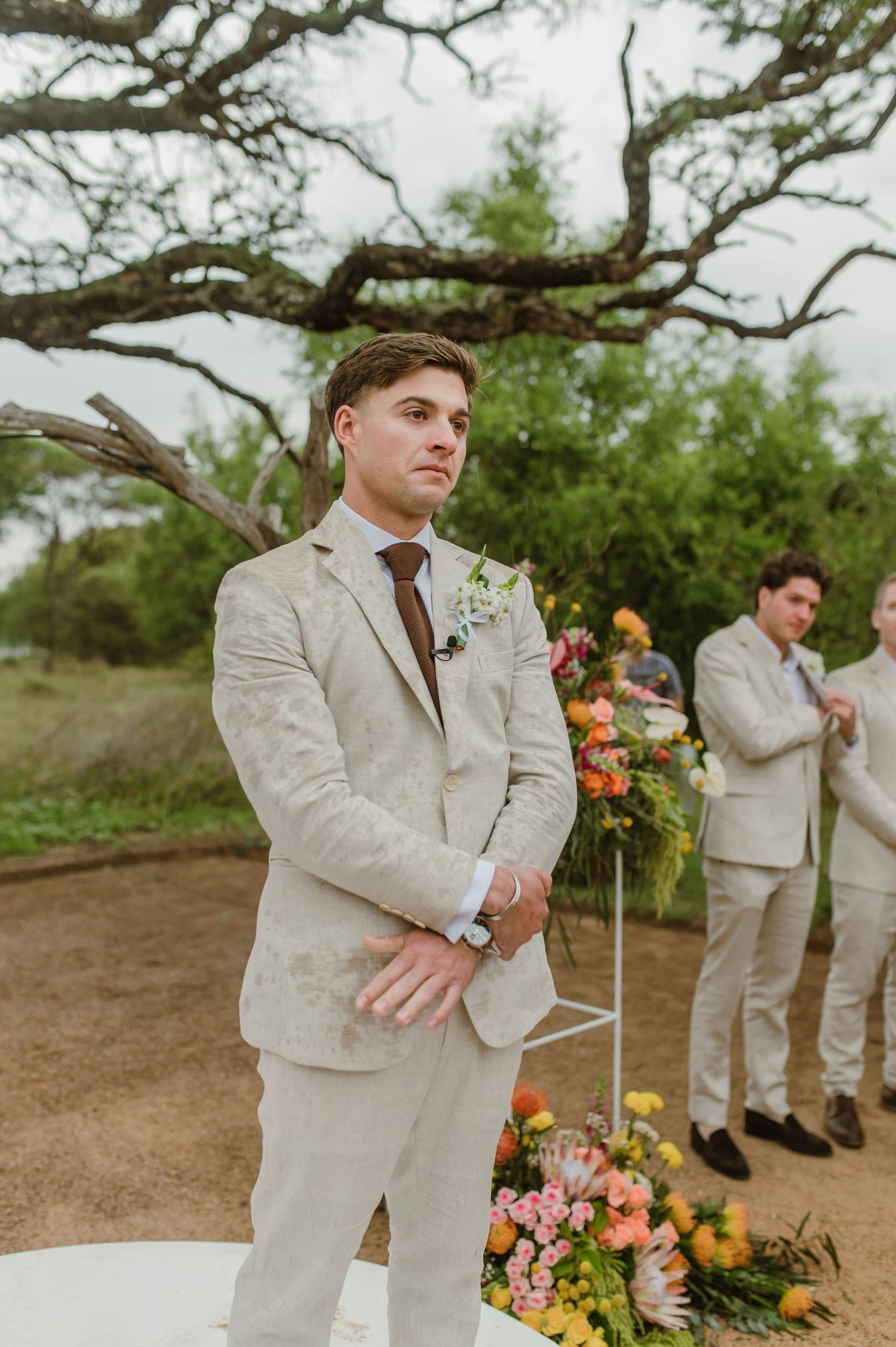 Wedding party members in beige suits pose near colorful floral arrangements outdoors.