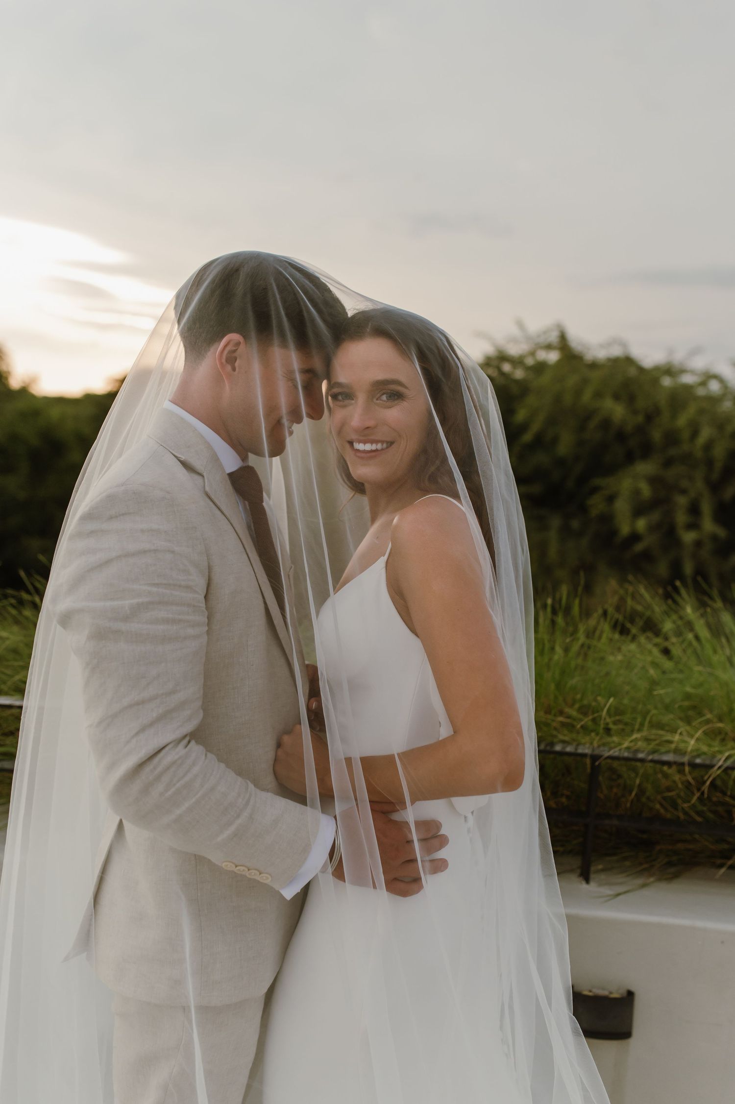 Romantic wedding couple shares an intimate moment under a flowing veil against a scenic outdoor backdrop.