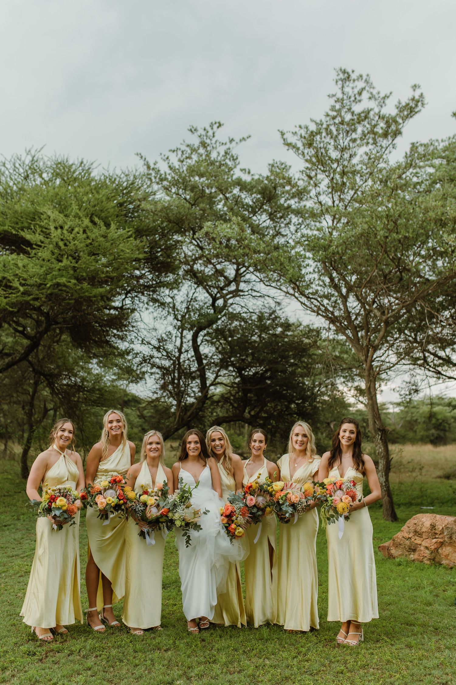 Wedding party in pale yellow dresses poses together on grassy lawn with trees in background holding colorful bouquets.