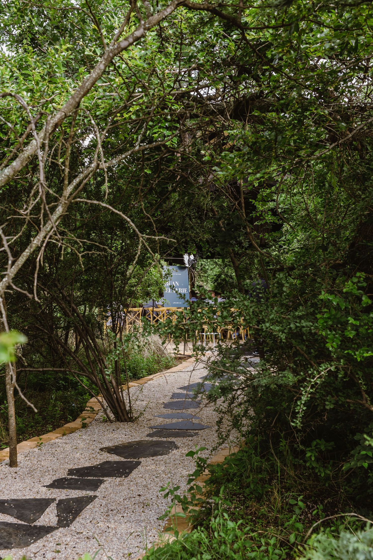 Shaded garden pathway with gravel leading through lush green trees.