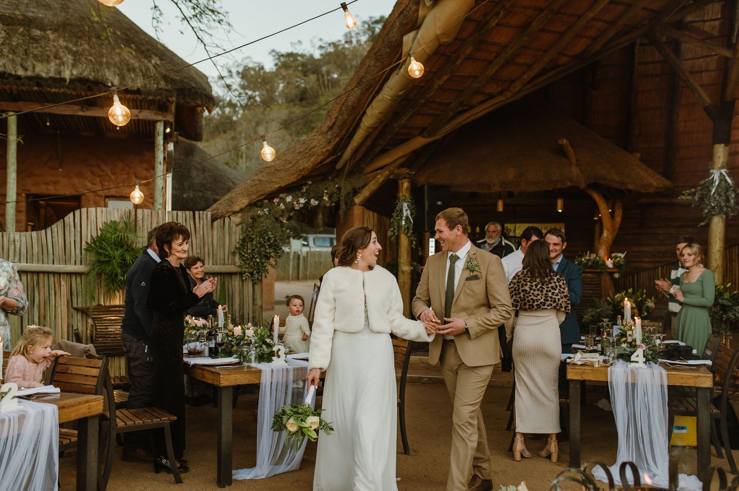 Indoor wedding reception with guests dining at decorated tables under wooden beams and string lights.