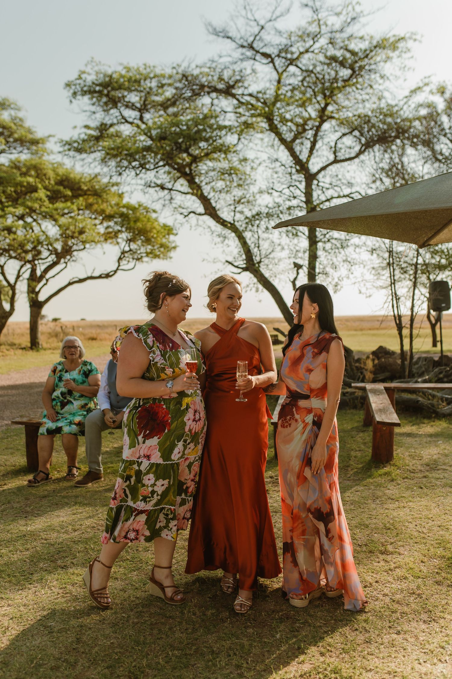 Wedding party in colorful dresses mingles outdoors under trees.